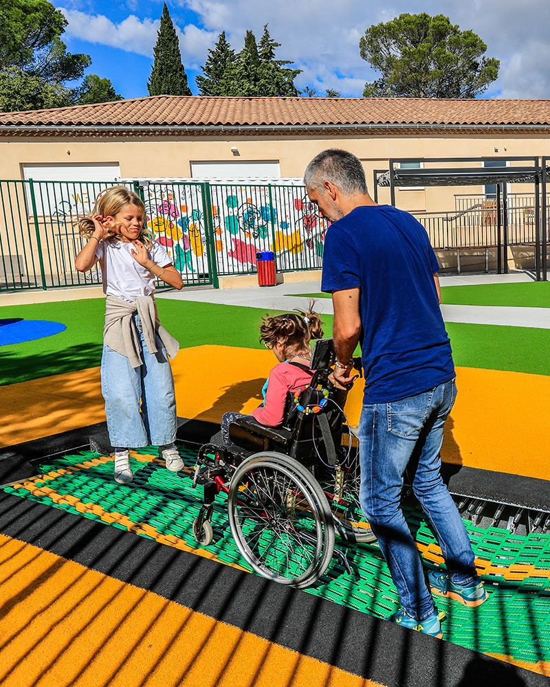 A parent takes a wheelchair user and another child onto an in ground trampoline for jumping on.